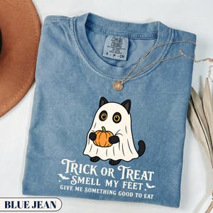 A funny Halloween graphic t-shirt featuring a cute black cat wearing a classic ghost sheet costume and holding a small pumpkin. The vintage style text reads TRICK OR TREAT SMELL MY FEET GIVE ME SOMETHING GOOD TO EAT with small flying bats. Perfect retro spooky season apparel and a funny cat lover gift.