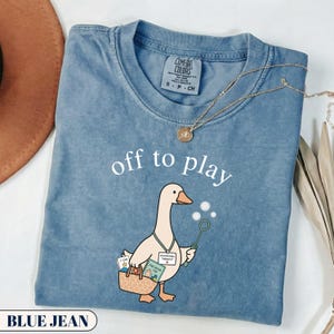 Può includere: Una t-shirt color blu jeans con la grafica di un'oca che tiene in mano una bacchetta per bolle di sapone e una borsa con la scritta "off to play". L'oca indossa un cordino con un badge "OCCUPATIONAL THERAPIST". La t-shirt ha un girocollo.