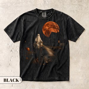 May include: A black short-sleeve t-shirt featuring a graphic print of a ghost riding a bicycle on a dark path. The scene includes a large orange full moon, bare trees, a pumpkin, and a house silhouette, creating a spooky nighttime atmosphere.