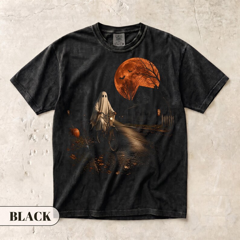 May include: A black short-sleeve t-shirt featuring a graphic print of a ghost riding a bicycle on a dark path. The scene includes a large orange full moon, bare trees, a pumpkin, and a house silhouette, creating a spooky nighttime atmosphere.