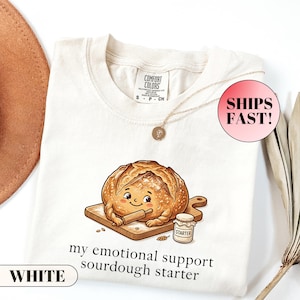 May include: An off-white crew neck t-shirt featuring a graphic of a cartoon sourdough loaf holding a rolling pin on a wooden board next to a jar labelled STARTER. The text below reads my emotional support sourdough starter.