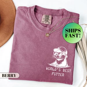 Pode incluir: Camiseta cor de berry com um gr&aacute;fico de uma lontra usando &oacute;culos de sol e chap&eacute;u, com o texto "WORLD'S BEST FOTTER". A camisa tem gola redonda e uma pequena etiqueta. Um colar de ouro est&aacute; drapeado sobre a camisa.