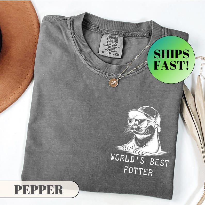 Pode incluir: Uma camiseta cinza Comfort Colors com um gr&aacute;fico de uma lontra usando &oacute;culos de sol e um chap&eacute;u. O texto "WORLD'S BEST FOTTER" est&aacute; abaixo da lontra. Um colar de prata e um c&iacute;rculo verde com as palavras "SHIPS FAST!" tamb&eacute;m s&atilde;o vis&iacute;veis.