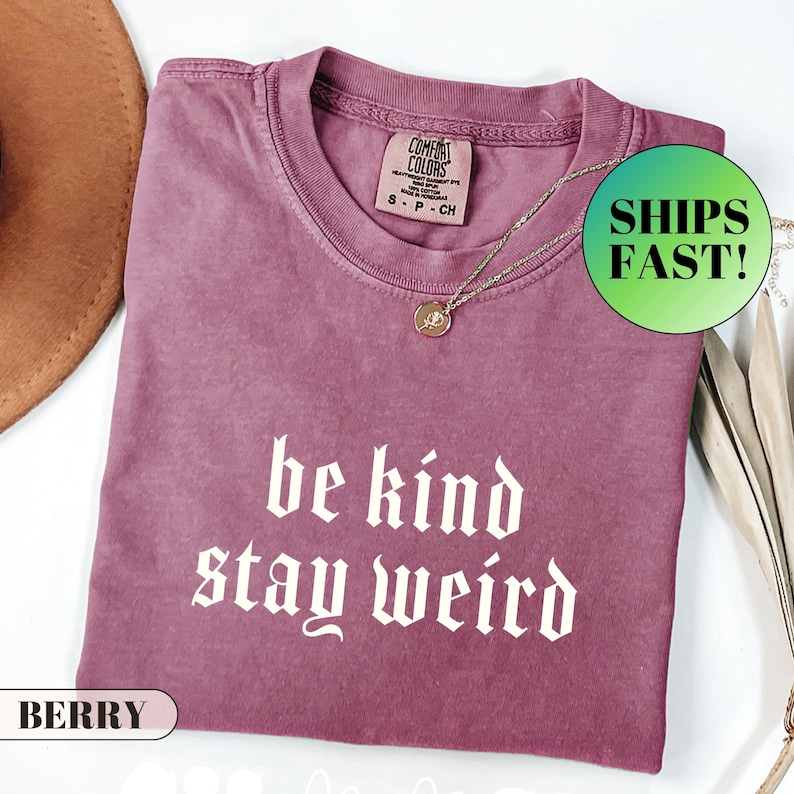 K&ouml;nnte beinhalten: Beerenfarbenes T-Shirt mit dem wei&szlig;en Schriftzug &bdquo;be kind stay weird&ldquo;. Das Shirt hat einen Rundhalsausschnitt und kurze &Auml;rmel. Eine goldfarbene Halskette mit kleinem Anh&auml;nger befindet sich auf dem Shirt. Das Shirt ist gefaltet und hat ein Etikett mit der Aufschrift &bdquo;Comfort Colors&ldquo;.