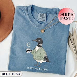 May include: A blue jean coloured t-shirt featuring a graphic of a loon wearing a light green cardigan and holding a steaming mug of coffee. The text below the bird reads, leave me a loon. The shirt has a crew neckline and a Comfort Colors brand label.