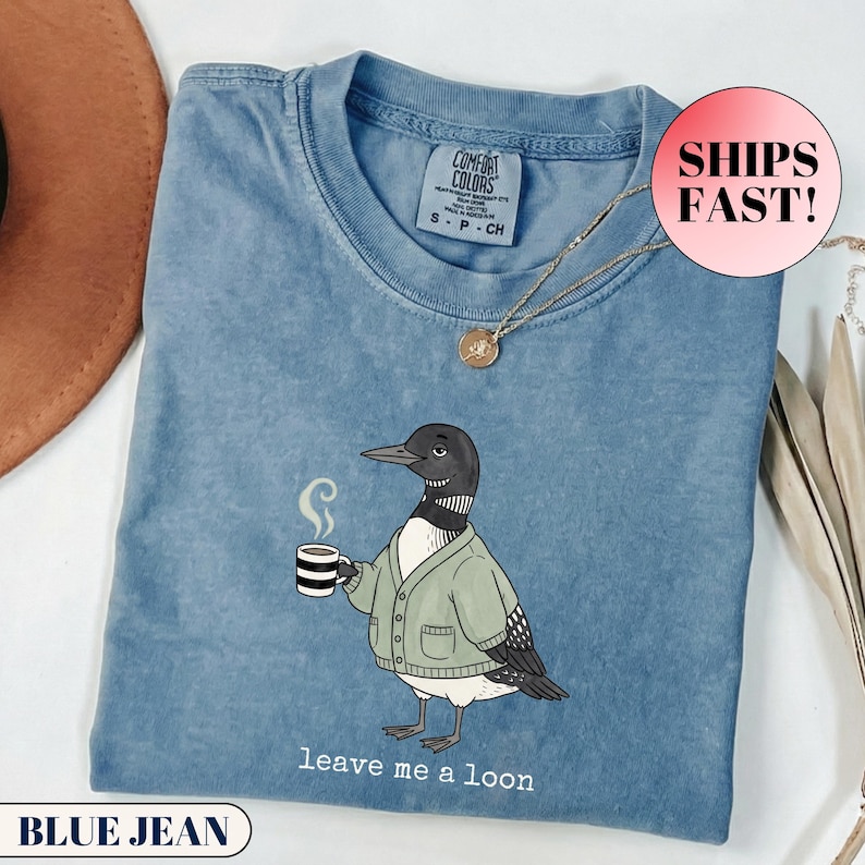 Flat lay of a folded Comfort Colors t-shirt. The minimalist graphic features a cartoon loon bird wearing a cozy green cardigan and holding a steaming mug of coffee. Below the bird, playful typewriter text reads "leave me a loon". Introvert bird meme shirt, sarcastic animal graphic tee, and funny coffee lover apparel.