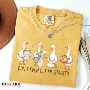 May include: A mustard yellow t-shirt featuring a graphic of four white geese engaged in baking activities, including holding jars and a loaf of bread. The text "DON'T EVEN GET ME STARTED" is printed below the geese. The shirt has a crew neckline.