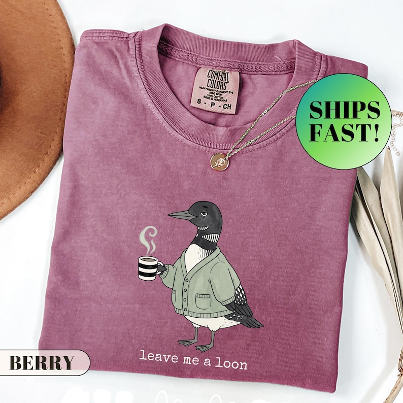 May include: A berry-colored crew neck t-shirt featuring a graphic of a loon wearing a light green cardigan and holding a steaming mug of coffee. The text below the graphic reads "leave me a loon". The shirt has a Comfort Colors brand label.