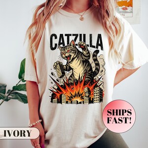 May include: Ivory-colored t-shirt featuring a graphic of a cat labeled "Catzilla" destroying buildings. The cat is gray and brown with red eyes, set against an explosion of orange and yellow. The shirt has a relaxed fit.