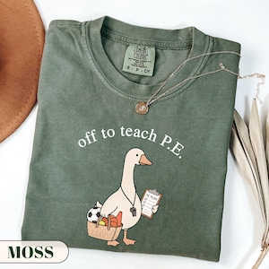 Op de afbeelding: Een mosgroen t-shirt met ronde hals en een afbeelding van een witte gans die een mand met een voetbal en basketbal vasthoudt, een fluitje draagt en een klembord met de tekst PE PLAN vasthoudt. De tekst erboven luidt off to teach P.E.