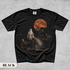 A vintage Halloween graphic t-shirt featuring a spooky ghost in a classic white sheet costume riding a bicycle down a dark street. The nostalgic night scene includes a glowing orange full moon, flying bats, a creepy house, and a carved pumpkin. Perfect retro fall apparel, spooky season trick or treat outfit, and nostalgic autumn clothing.