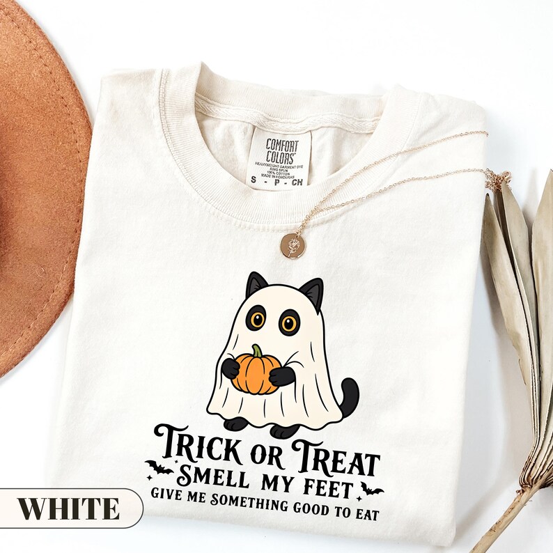 A funny Halloween graphic t-shirt featuring a cute black cat wearing a classic ghost sheet costume and holding a small pumpkin. The vintage style text reads TRICK OR TREAT SMELL MY FEET GIVE ME SOMETHING GOOD TO EAT with small flying bats. Perfect retro spooky season apparel and a funny cat lover gift.