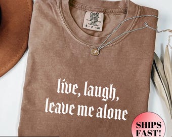 May include: Espresso-colored t-shirt with the phrase "live, laugh, leave me alone" in white gothic font. The shirt has a crew neck and short sleeves. A silver necklace with a pendant is draped across the shirt.