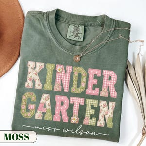 May include: A sage green short-sleeve t-shirt with a crew neckline and a Comfort Colors label. The front features the word KINDERGARTEN in large block letters with floral and gingham patterns, plus the cursive text miss wilson below it.