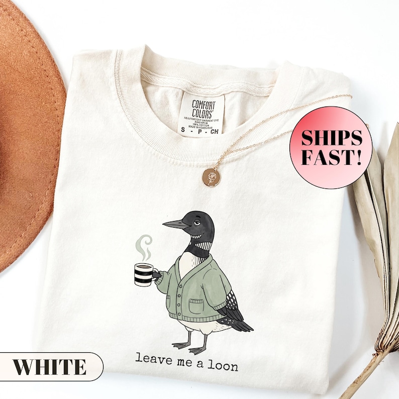 Flat lay of a folded Comfort Colors t-shirt. The minimalist graphic features a cartoon loon bird wearing a cozy green cardigan and holding a steaming mug of coffee. Below the bird, playful typewriter text reads "leave me a loon". Introvert bird meme shirt, sarcastic animal graphic tee, and funny coffee lover apparel.
