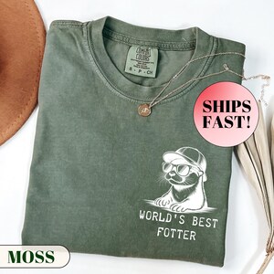 Pode incluir: Uma camiseta verde musgo com um gr&aacute;fico de uma lontra usando &oacute;culos de sol e bon&eacute;, com o texto "WORLD'S BEST FOTTER". A camisa tem gola redonda e uma etiqueta pequena. Um colar de ouro com um pingente est&aacute; sobre a camisa.