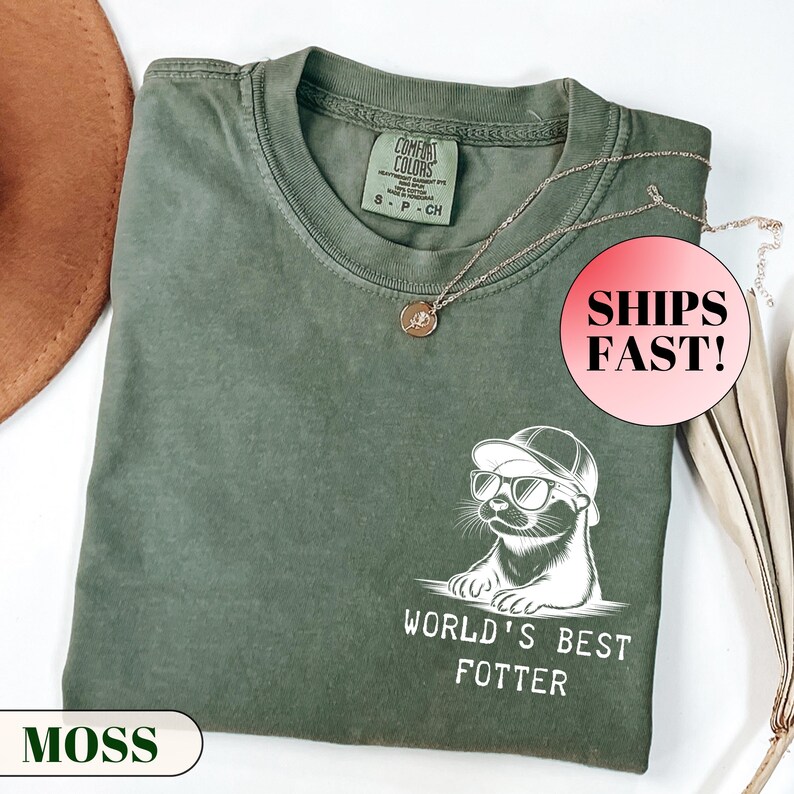 Pode incluir: Uma camiseta verde musgo com um gr&aacute;fico de uma lontra usando &oacute;culos de sol e bon&eacute;, com o texto "WORLD'S BEST FOTTER". A camisa tem gola redonda e uma etiqueta pequena. Um colar de ouro com um pingente est&aacute; sobre a camisa.