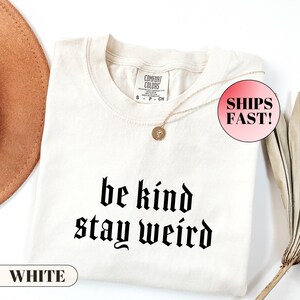 K&ouml;nnte beinhalten: Cremefarbenes T-Shirt mit dem Schriftzug "be kind stay weird" in schwarzer gotischer Schrift. Das Shirt hat einen Rundhalsausschnitt und kurze &Auml;rmel. Eine goldene Halskette mit Anh&auml;nger liegt auf dem Shirt. Das Wort "WHITE" ist auf einem wei&szlig;en Rechteck aufgedruckt.