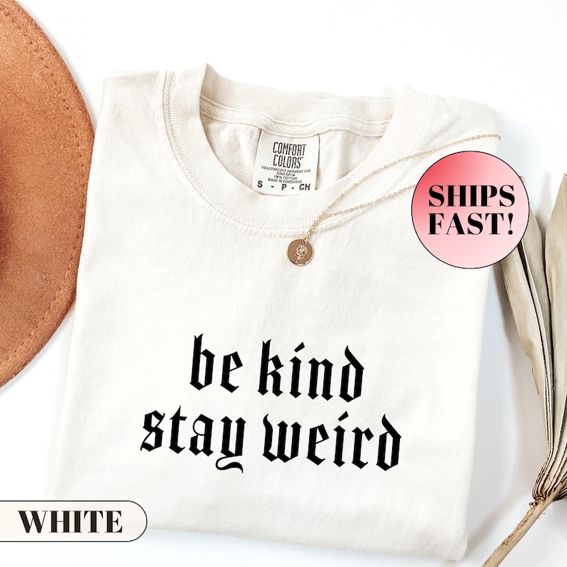 K&ouml;nnte beinhalten: Cremefarbenes T-Shirt mit dem Schriftzug "be kind stay weird" in schwarzer gotischer Schrift. Das Shirt hat einen Rundhalsausschnitt und kurze &Auml;rmel. Eine goldene Halskette mit Anh&auml;nger liegt auf dem Shirt. Das Wort "WHITE" ist auf einem wei&szlig;en Rechteck aufgedruckt.