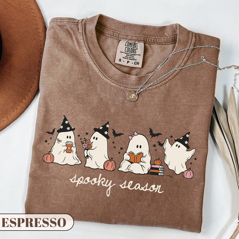 A cute pastel Halloween graphic t-shirt featuring four kawaii sheet ghosts wearing witch hats and bows, drinking coffee, holding flowers, and reading a book next to pumpkins and bats. The cursive text reads spooky season. Perfect cozy autumn apparel and bookish fall gift.