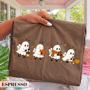 May include: A brown t-shirt featuring a graphic of four cartoon bees dressed as ghosts. The bees are depicted holding an acorn lantern, a maple leaf, a red apple, and small mushrooms, surrounded by scattered autumn leaves in orange and yellow.