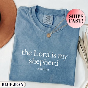 May include: A blue jean colored short-sleeve t-shirt with a crew neckline. The shirt features the white printed text "the Lord is my shepherd" followed by "psalm 23:1". A gold-toned pendant necklace rests on the collar area of the garment.