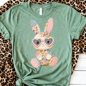 Könnte beinhalten: Salbeigrünes T-Shirt mit einem Patchwork-Hasen, der eine herzförmige Brille trägt. Der Hase hat Blumen- und Vichy-Muster in Ohren, Körper und Füßen. Die Brille ist mit Perlen verziert. Das T-Shirt ist aus weichem Material.