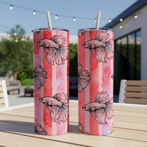 May include: Two stainless steel tumblers with a red and white striped watercolor design. Each tumbler features a repeating pattern of pink betta fish illustrations. The tumblers have white straws.