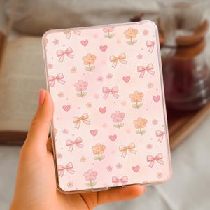 Op de afbeelding: Een doorzichtige tablethoes met een herhaald patroon van roze strikken, bloemen en hartjes op een lichtroze achtergrond. De hoes wordt in een hand gehouden, waarbij de details van het ontwerp worden benadrukt.