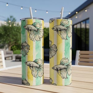 May include: Two stainless steel tumblers featuring a betta fish design. The fish are illustrated in black and white, set against vertical yellow and green watercolor stripes. Each tumbler has a white straw.