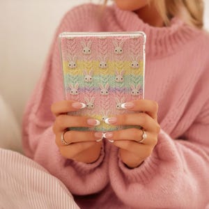 May include: A clear phone case featuring a pastel rainbow knit pattern with repeating white bunny faces. The case is held in front of a pink sweater. The case is a cute and whimsical accessory.