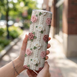 Op de afbeelding: Een roestvrijstalen beker met een crèmekleurig gebreid patroon en geborduurde rozen in roze, wit en bordeauxrood. Het bloemmotief is in een herhalend patroon gerangschikt. De beker is cilindrisch en lijkt geïsoleerd.