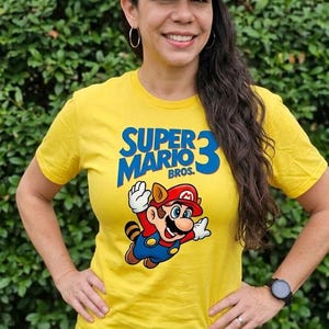 May include: A yellow Super Mario Bros. 3 t-shirt featuring a graphic of Mario in mid-air. The person is wearing a navy blue baseball cap with the number 46 and the words "The Doctor" on it, along with denim shorts.