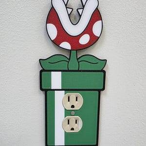 May include: A decorative wall outlet cover depicting a Piranha Plant from the Super Mario video game series. The cover has a green pot with white stripes and two electrical outlets. The Piranha Plant has a red head with white spots and a white mouth.