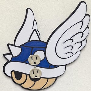 May include: A decorative wall outlet cover featuring a blue, white, and tan cartoon design with two electrical outlets. The design includes wings and a spiky shell, with black outlines.
