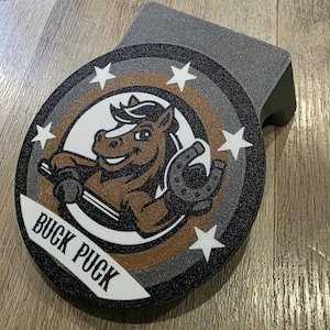 May include: A gray and black hockey puck holder with a cartoon horse graphic. The horse is brown and white, holding a horseshoe. The text "BUCK PUCK" is displayed on a white banner.