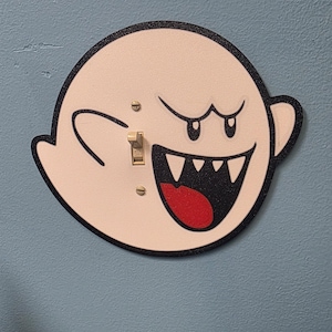 May include: A decorative light switch cover featuring a cartoon ghost character with a black outline, white body, and red mouth. The switch is a standard toggle style, and the cover is mounted on a blue wall.