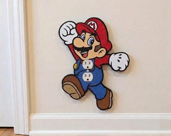 May include: A decorative wall plate cover shaped like the character Mario, featuring a red hat with an M, blue overalls, and white gloves. The design incorporates two white electrical outlet receptacles positioned directly on the character's torso.