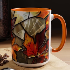 May include: A white ceramic mug with an orange handle and interior, featuring a stained-glass design of autumn leaves in shades of red, orange, and yellow. The mug sits on a wooden board with a macaron and a spoon.