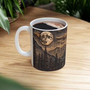 May include: White ceramic coffee mug with a black handle. The mug features a graphic of a full moon over a dark forest and mountain landscape. The moon is a light beige color and the forest and mountains are a dark brown color.