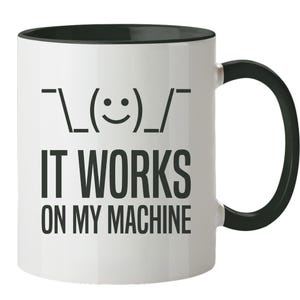 Op de afbeelding: Een witte keramische mok met een zwarte binnenkant en een zwart handvat. De voorzijde van de mok bevat een zwarte afbeelding van een schouderophalende emoticon boven de zwarte tekst IT WORKS ON MY MACHINE in een vet, schreefloos lettertype.