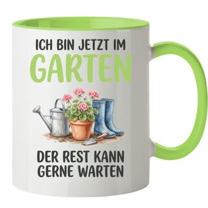 Op de afbeelding: Een witte keramische mok met een limoengroene binnenkant en handvat. De voorzijde toont een afbeelding van een zilveren gieter, een terracotta pot met roze bloemen, blauwe laarzen en een troffel. Tekst: ICH BIN JETZT IM GARTEN DER REST KANN GERNE WARTEN.