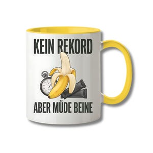 Könnte beinhalten: Ein weißer Keramikbecher mit gelbem Innenraum und gelbem Henkel, bedruckt mit einer Grafik einer geschälten Banane, einer silbernen Stoppuhr und zwei schwarzen rechteckigen Päckchen. Der Text "KEIN REKORD" steht oben und "ABER MÜDE BEINE" unten.