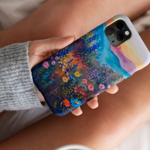 May include: A phone case with a watercolor design of a field of wildflowers and a mountain range in the background. The case is white with a clear back and a blue, purple, and pink floral design.