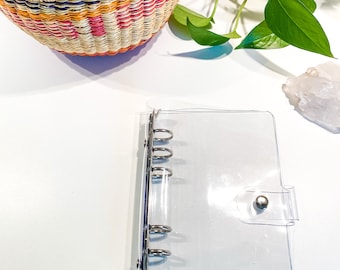 May include: A clear plastic binder with six silver rings and a silver snap closure. The binder is lying on a white surface.