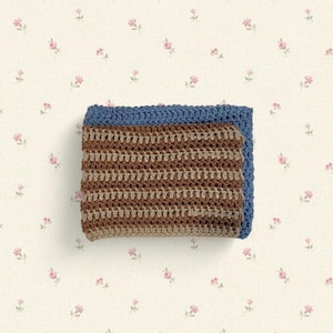 May include: A folded, handmade crochet blanket. The blanket features a blue border and a brown and tan striped pattern. The blanket is made of soft yarn and is perfect for snuggling. The background is a cream color with a pink floral pattern.