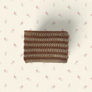 May include: A rectangular, crocheted item in shades of brown and tan. The crochet pattern features horizontal stripes. The item is displayed on a cream-colored background with a delicate floral pattern of pink flowers.