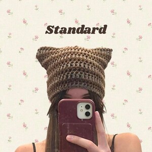 May include: A brown and tan crocheted cat ear hat. The hat is made of a textured yarn and has pointed ears at the top. The word "Standard" is printed above the hat.