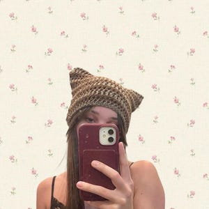 May include: A brown and tan crocheted cat ear hat. The hat is handmade with a textured stitch and features pointed ears. The person is holding a maroon phone in front of their face. The background is a cream-colored wallpaper with pink floral patterns.