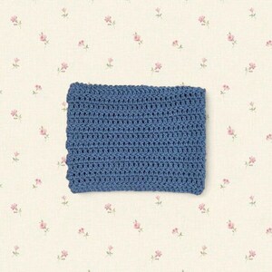 May include: A rectangular, crocheted item in a solid shade of blue. The handmade piece has a textured appearance, with visible stitches creating a pattern. The background is a cream color with a delicate floral print.